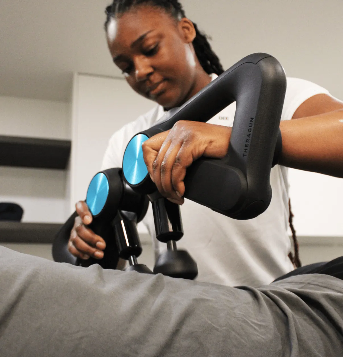 Person using Theragun massage device on another person's leg wearing gray pants.