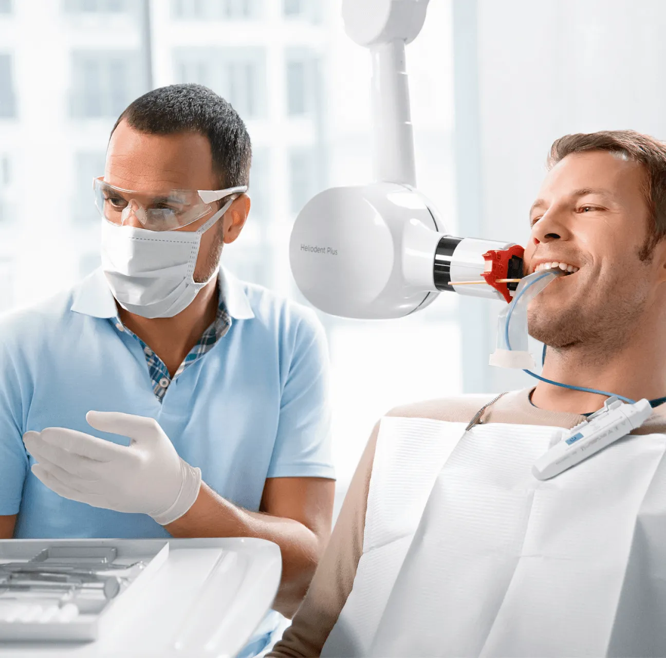 Dentist wearing safety glasses and mask preparing to use a dental X-ray machine on a smiling male patient with a bite block in his mouth.