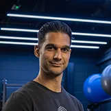 Man with dark hair and a slight smile standing in a gym with blue exercise balls in the background.