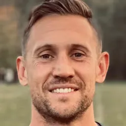 Close-up portrait of a smiling man with short brown hair and light facial hair against a blurred outdoor background.