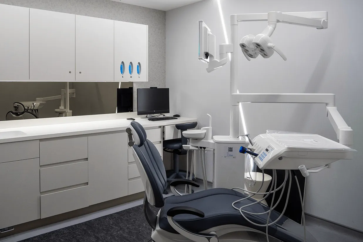 Modern dental clinic room with a dental chair, overhead dental light, computer workstation, and storage cabinets.