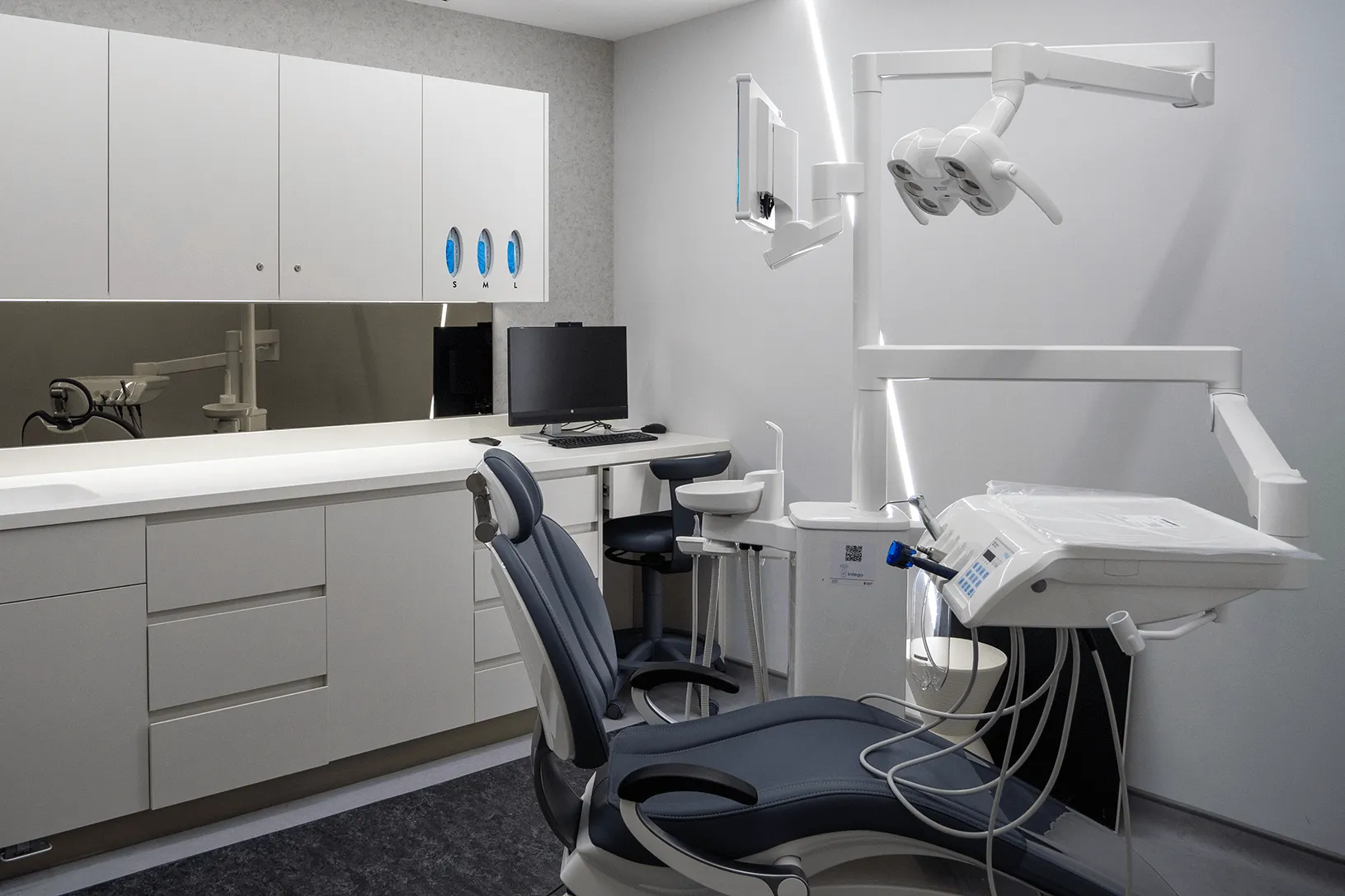 Modern dental examination room with dental chair, overhead light, monitor, and white cabinetry.