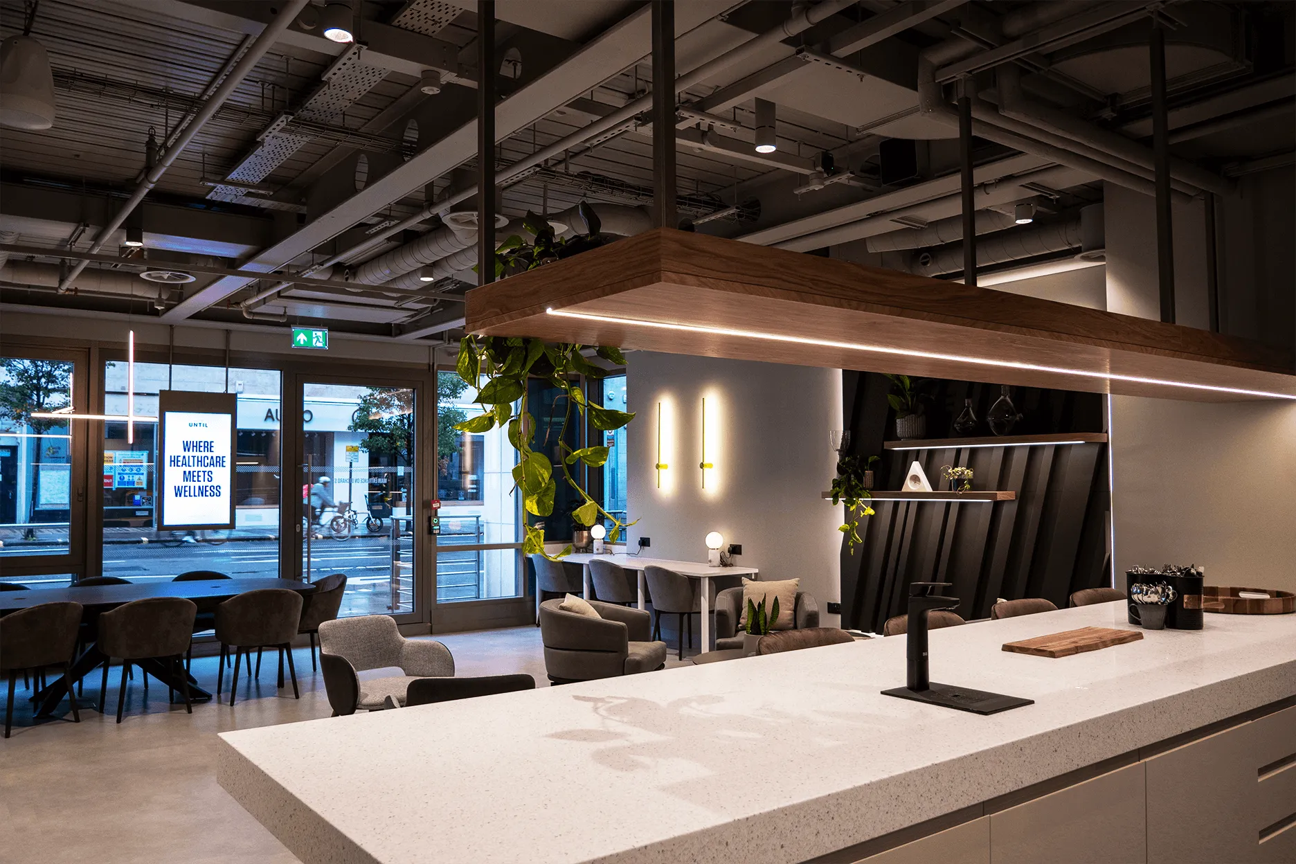 Modern wellness lounge with large white countertop island, gray chairs, plants, and a sign reading 'Where healthcare meets wellness' visible through glass doors.
