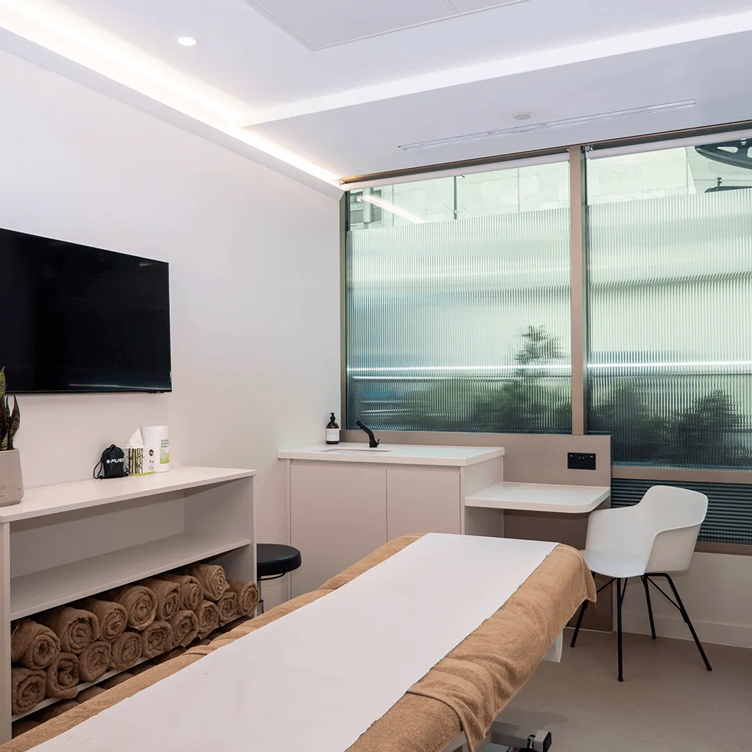 Modern massage room with treatment table covered in brown towels, white chair, sink, and soft lighting.
