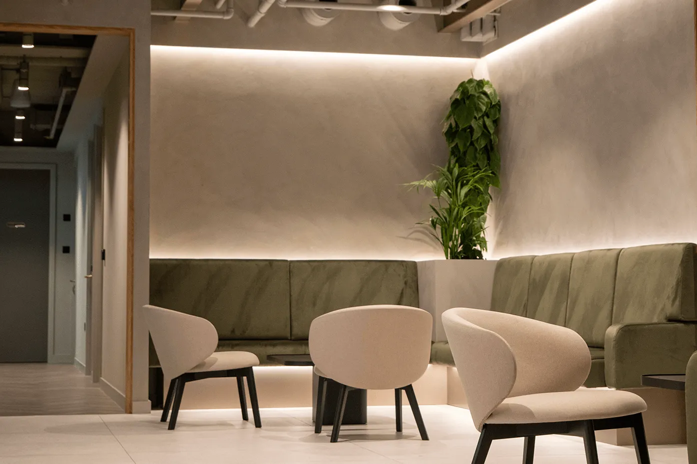 Modern reception area with beige chairs, a green upholstered sofa, ambient lighting, and tall plants in the corner.