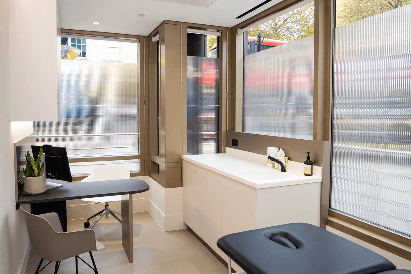 Modern medical examination room with frosted glass windows, black exam table, white cabinetry with sink, and a desk with a computer and chairs.
