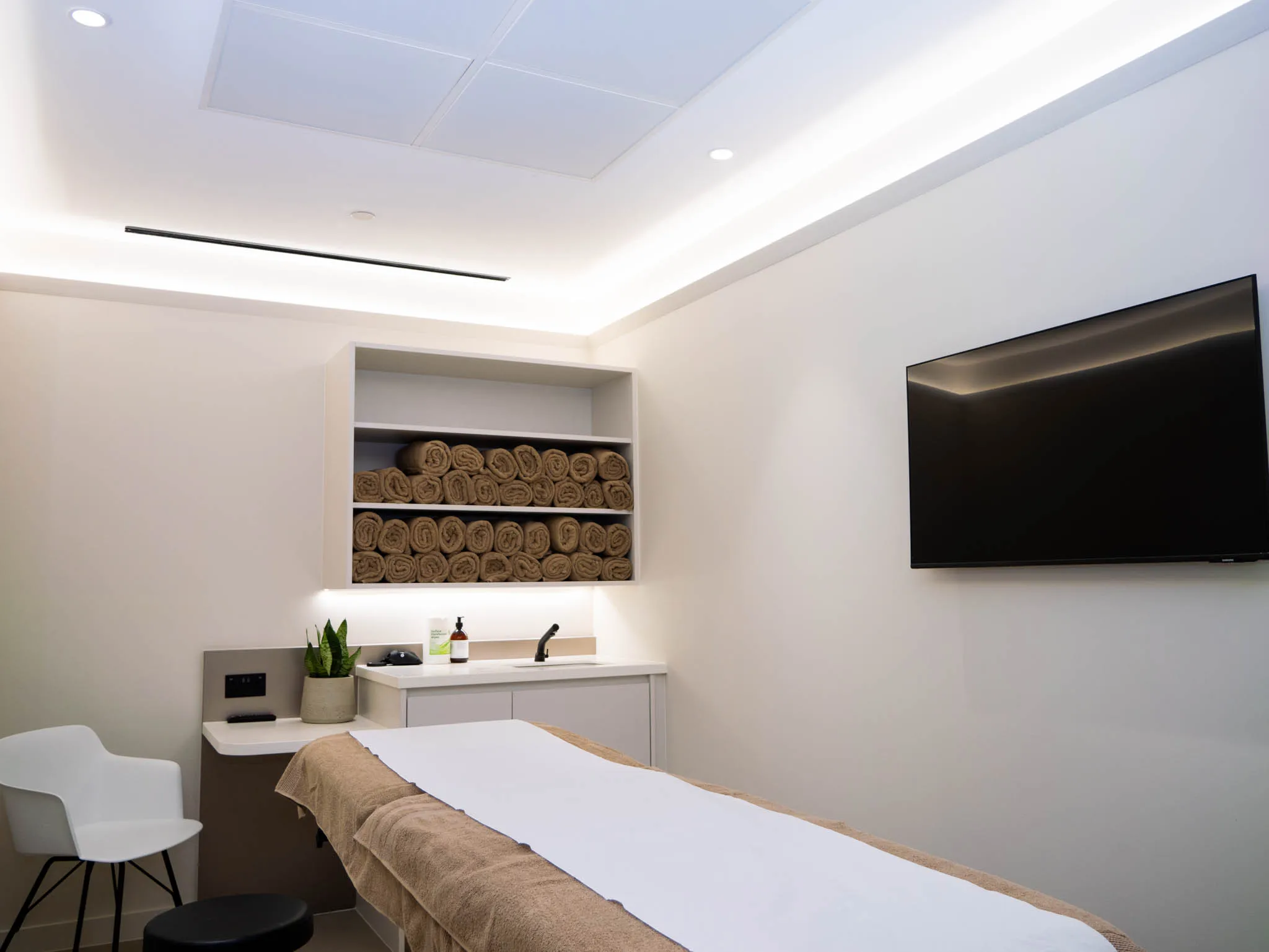 Minimalist spa room with massage table covered in brown towels, white chair, mounted TV, and stacked towels on shelves.