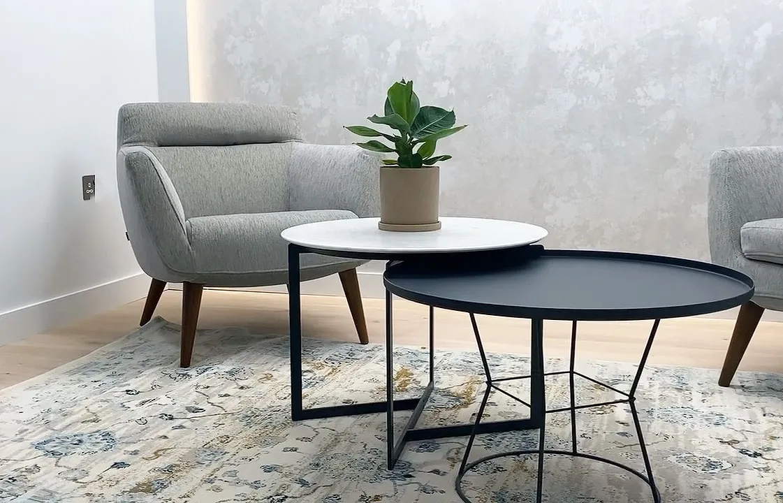 Modern living room with two gray armchairs, nested round coffee tables, and a small potted plant on a patterned rug.