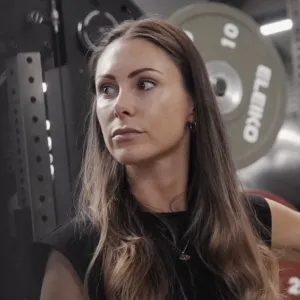 Woman with long brown hair standing in a gym with weight plates in the background.