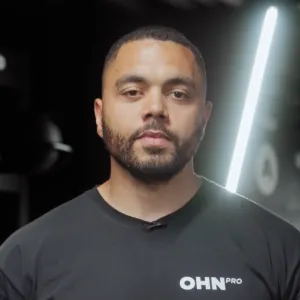 Man with short hair and beard wearing a black OHN Pro shirt against a dark blurred background with bright vertical light.