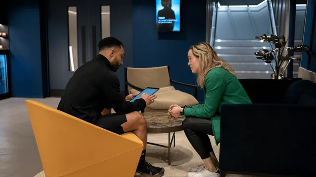 Two people sitting across from each other in a modern lounge area, engaged in conversation with a tablet device.