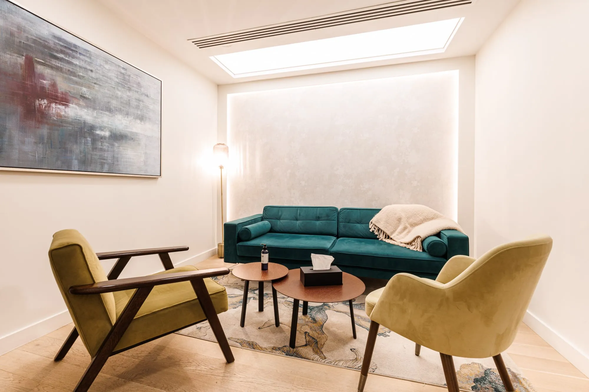 Modern living room with teal couch, beige armchairs, two round wooden coffee tables, and abstract wall art.
