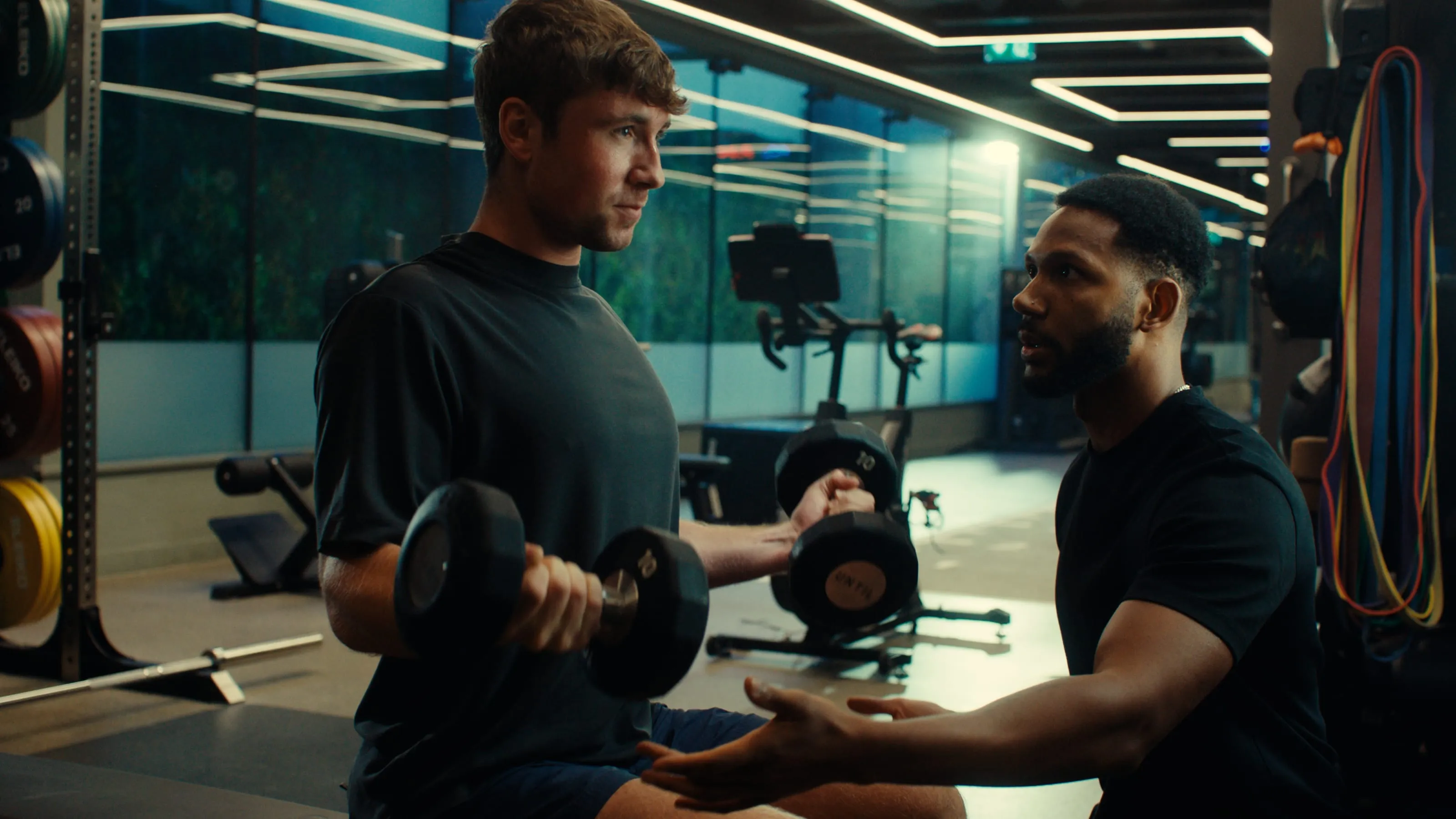 A man lifting dumbbells is coached by a trainer in a gym with exercise equipment and colorful resistance bands.