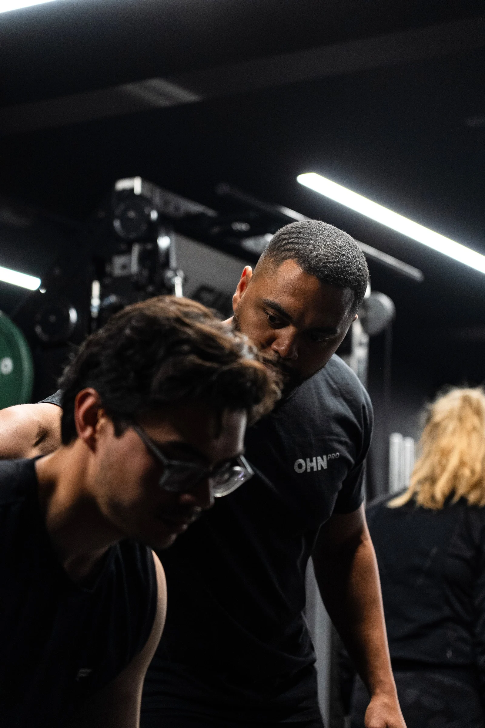 A fitness coach wearing a black OHN Pro shirt guiding a man with glasses during a workout in a gym.