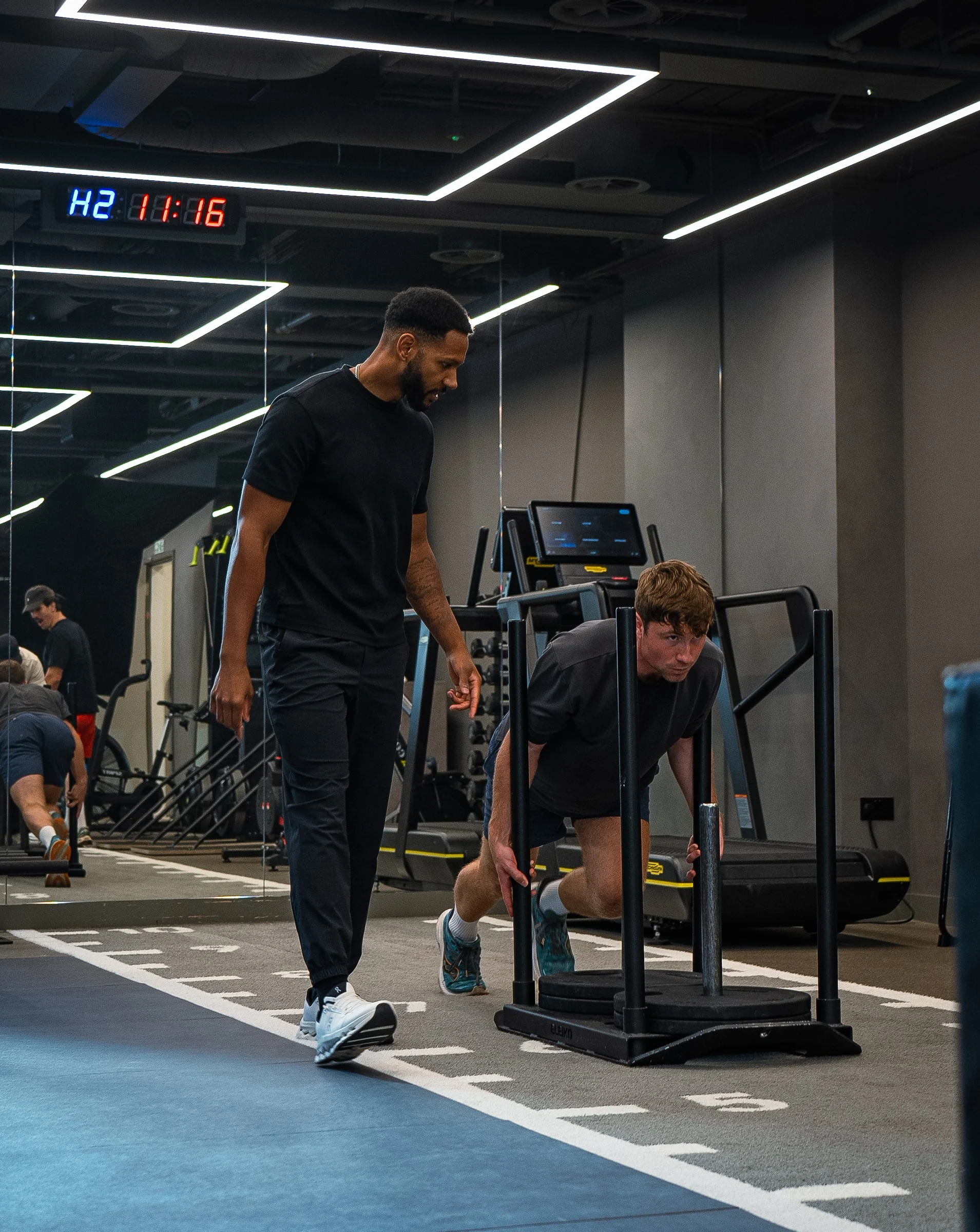 Man in black shirt coaching another man pushing a weighted sled on a gym track.