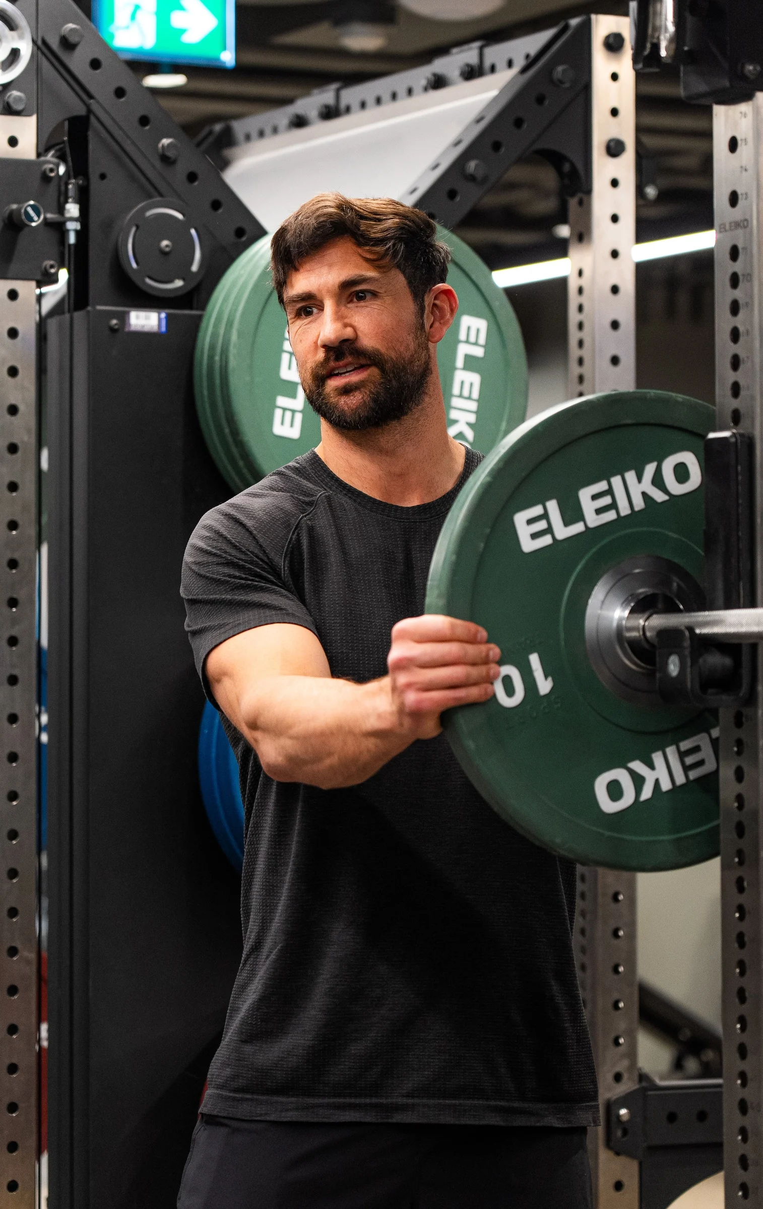 Man in black workout shirt loading a green Eleiko 10 kg weight plate onto a barbell in a gym.