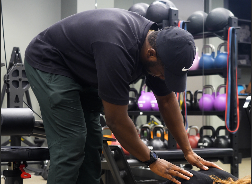Fitness trainer wearing a black cap and shirt assisting a person lying down for workout in a gym with kettlebells in the background.