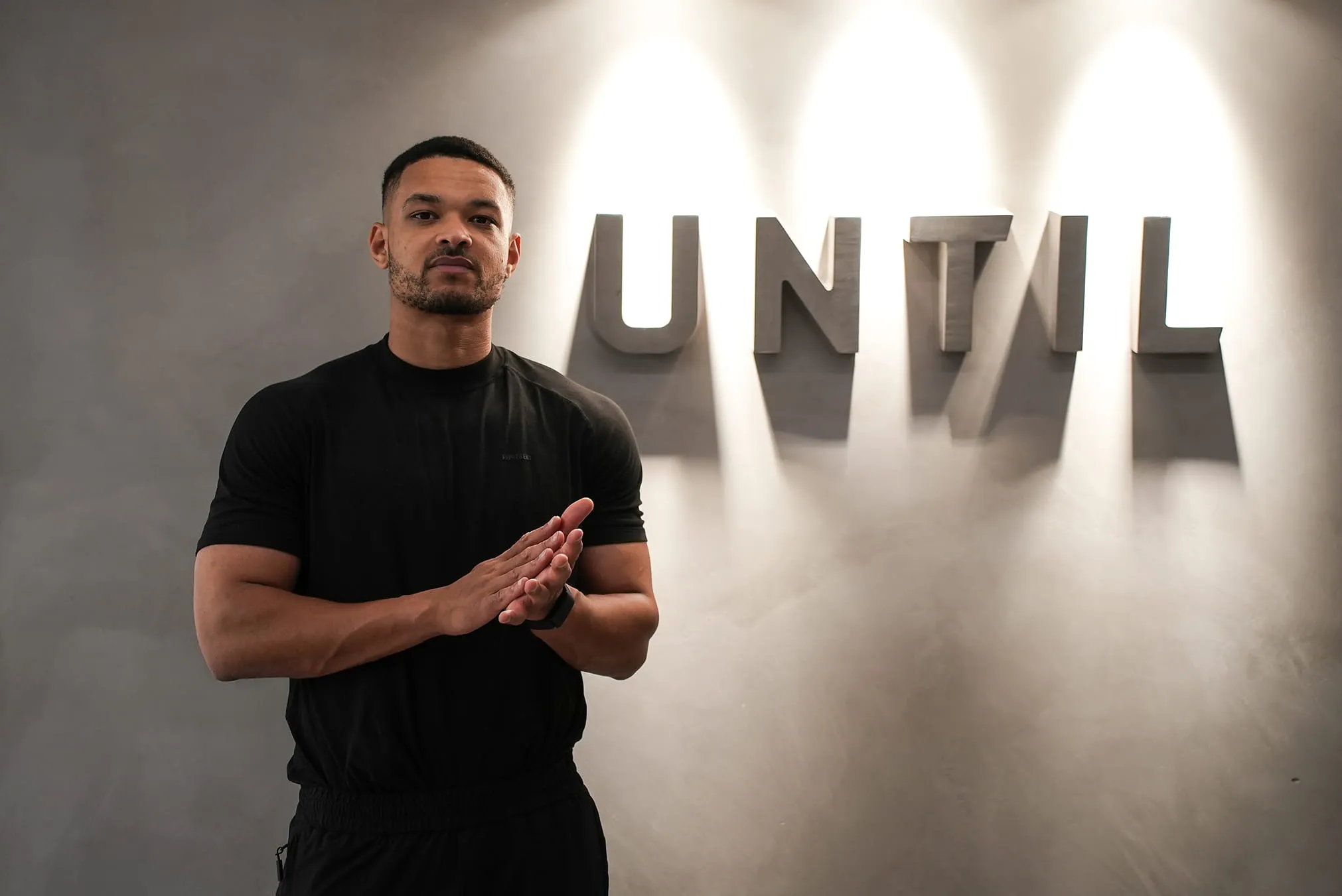 Man in black shirt standing with hands clasped in front of a wall displaying the word UNTIL in large letters.