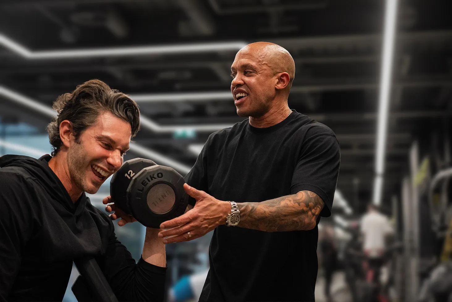 Trainer assisting a smiling man lifting a 12 kg dumbbell in a gym setting.