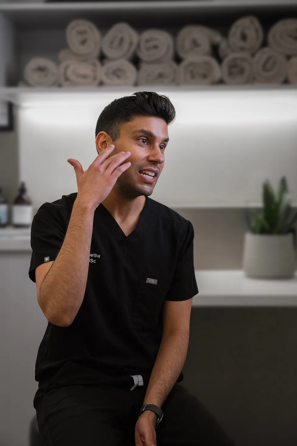 Man in black scrubs gesturing near his face, sitting in a room with rolled towels and a potted plant in the background.