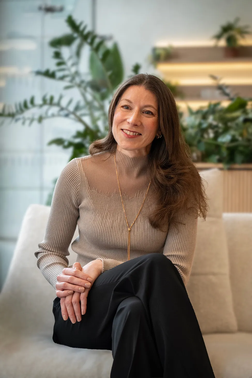 Smiling woman with long brown hair in a beige ribbed sweater and black pants sitting on a sofa with plants in the background.