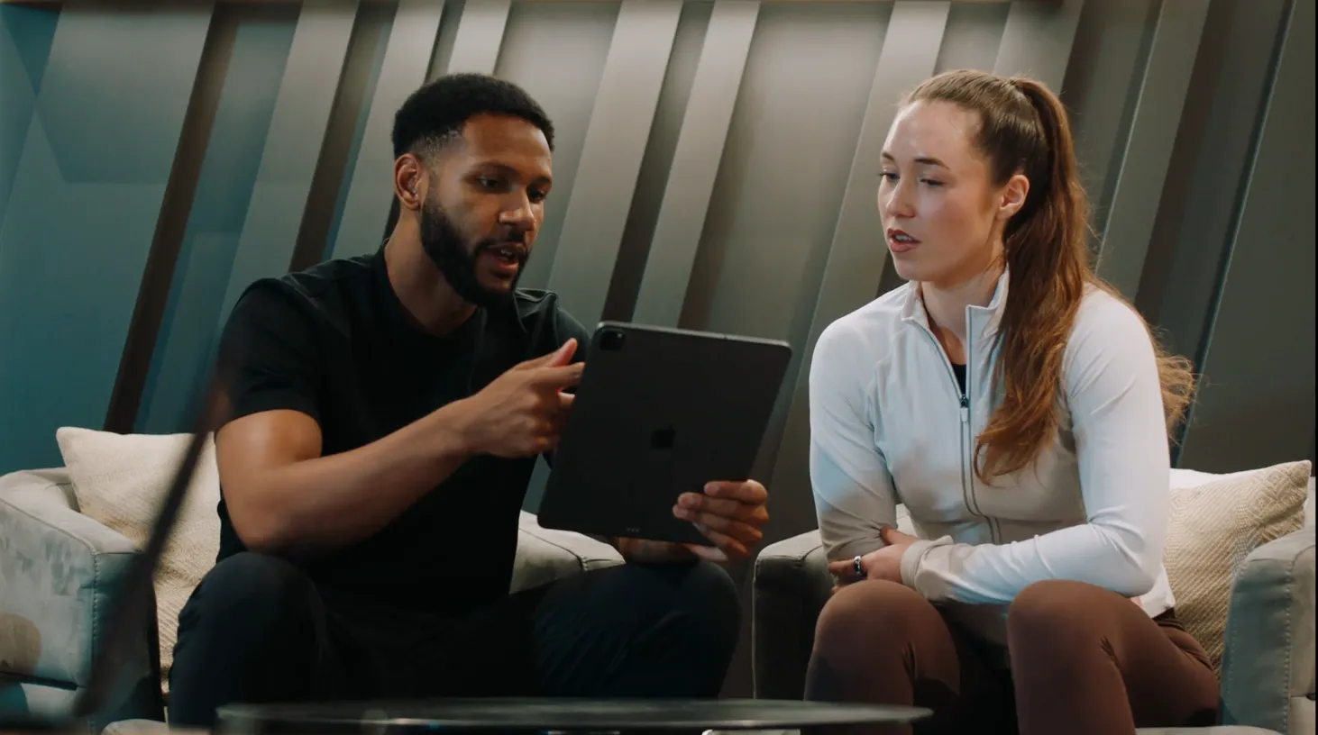 A man and a woman sitting on couches, looking at a tablet together in a modern room.