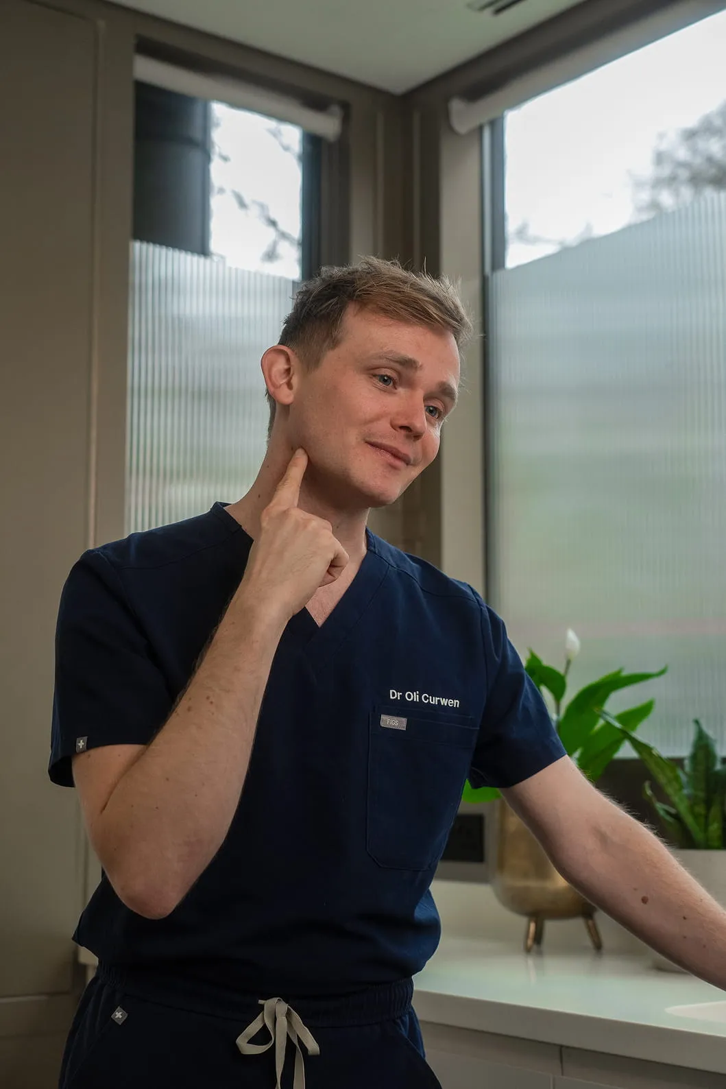 Doctor in navy blue scrubs pointing to the side of his neck in a clinical setting with plants and frosted windows behind.