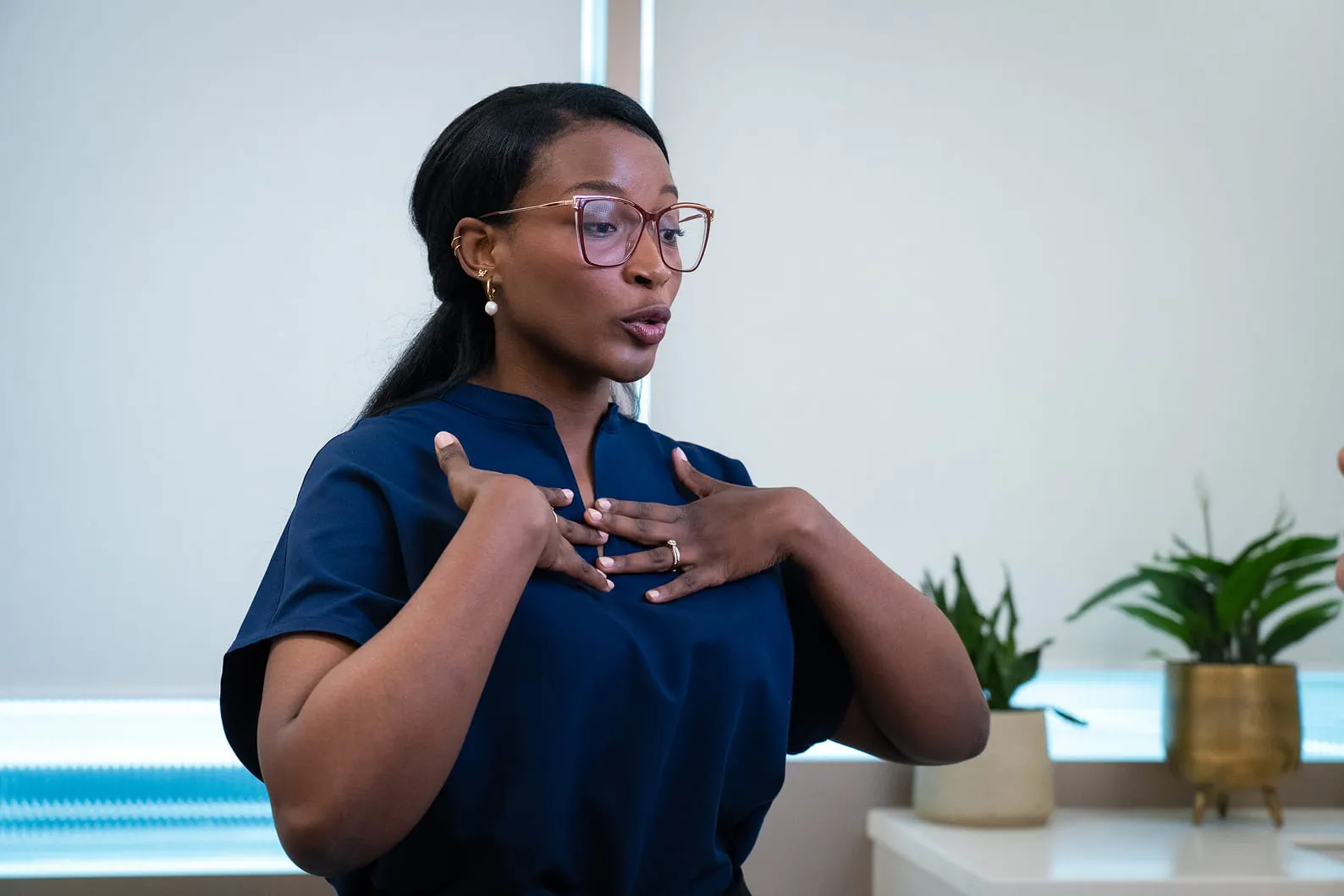 Woman in glasses wearing a navy blue top speaks with hands on her chest indoors near potted plants.