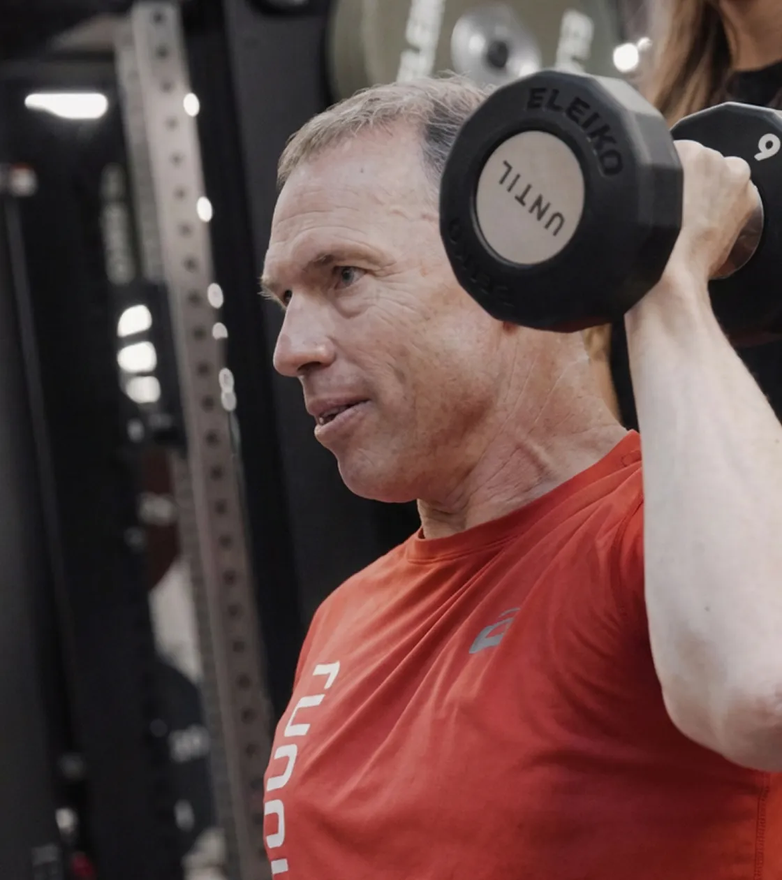 Middle-aged man in a red shirt lifting a black dumbbell in a gym.
