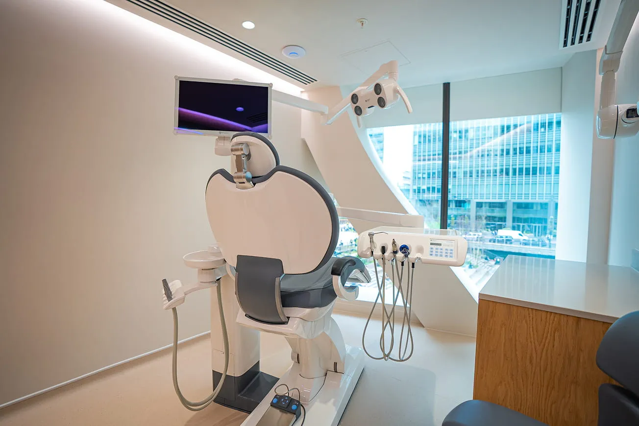 Modern dental chair with attached dental tools and overhead light in a bright clinic room with large window showing an urban building outside.