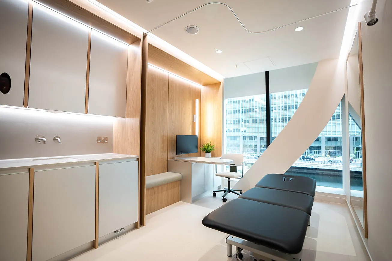 Modern medical examination room with black examination table, desk with computer, and large window showing city buildings.