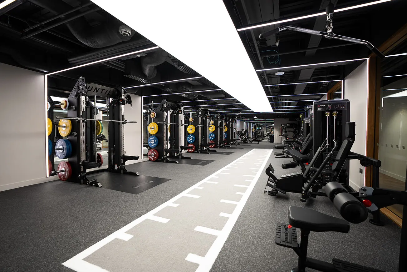 Modern gym interior with multiple squat racks holding colorful weight plates and various strength training machines along a mirrored wall.