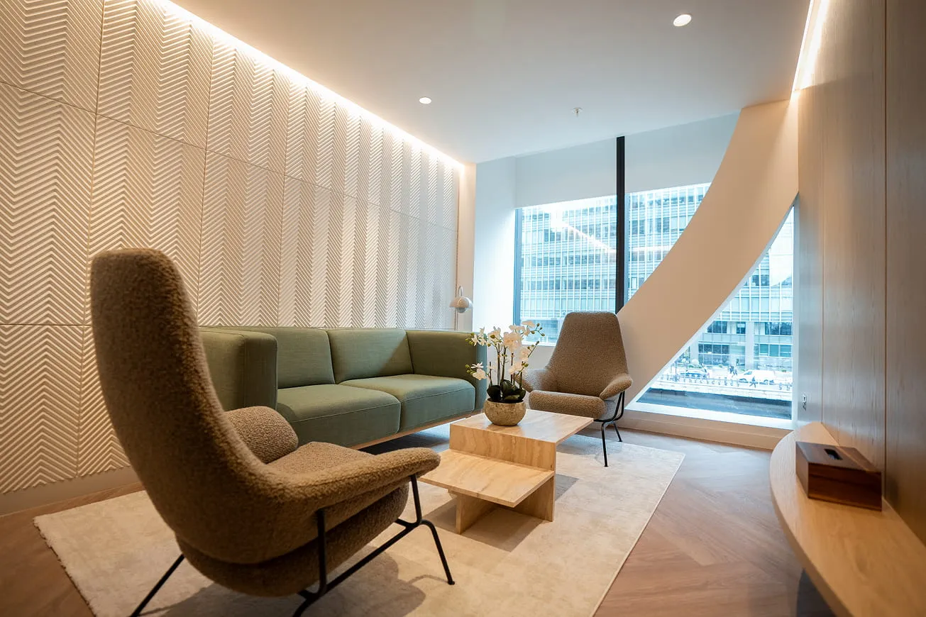 Modern office lounge with textured wall, green sofa, two beige armchairs, a light wood coffee table with flowers, and large window overlooking city buildings.