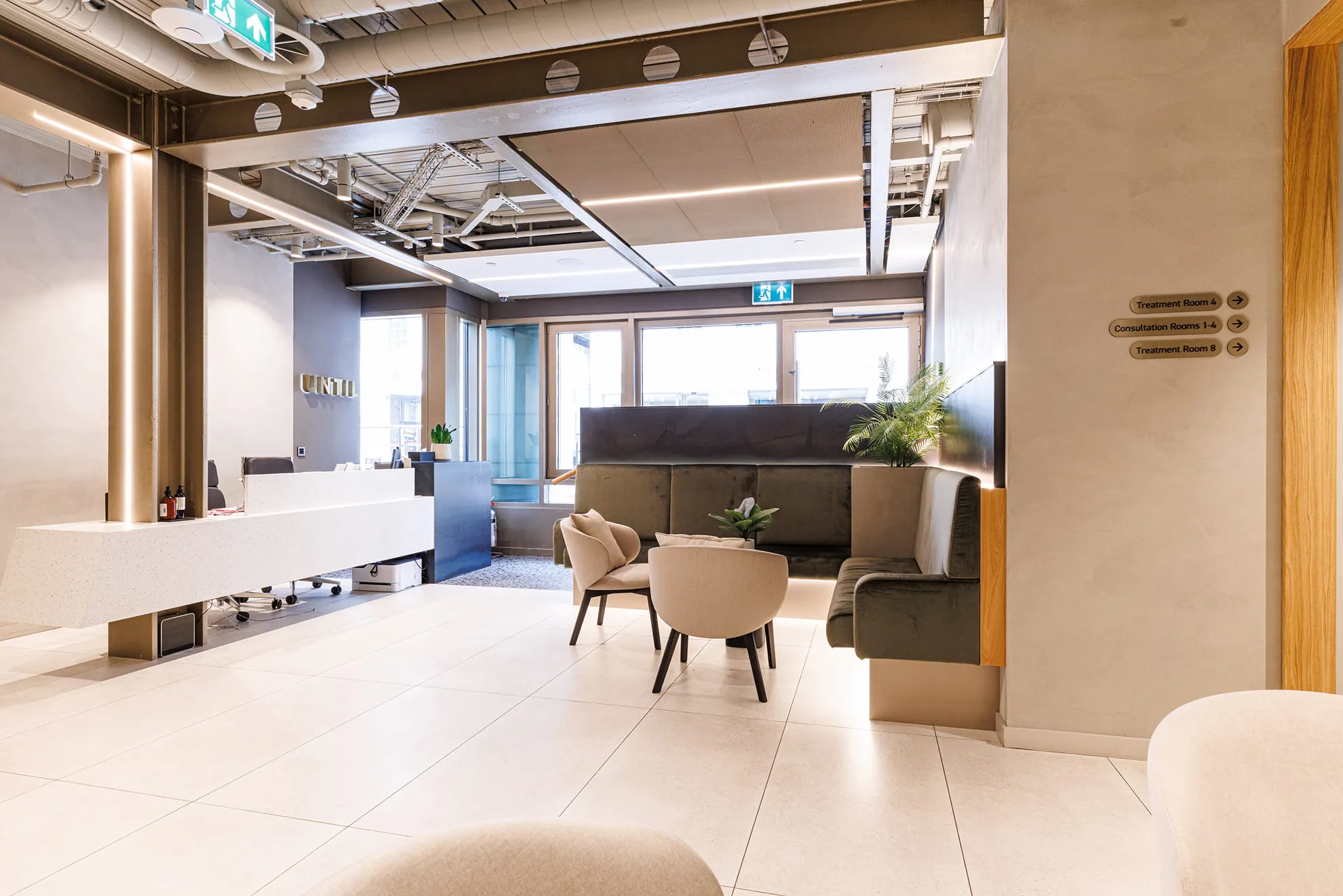 Modern waiting area with beige chairs, green cushioned bench, plants, reception desk, and directional signs.