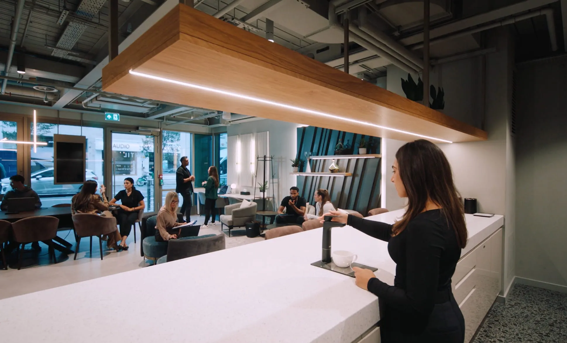 Woman making coffee at counter in modern office lounge where people are sitting and chatting.