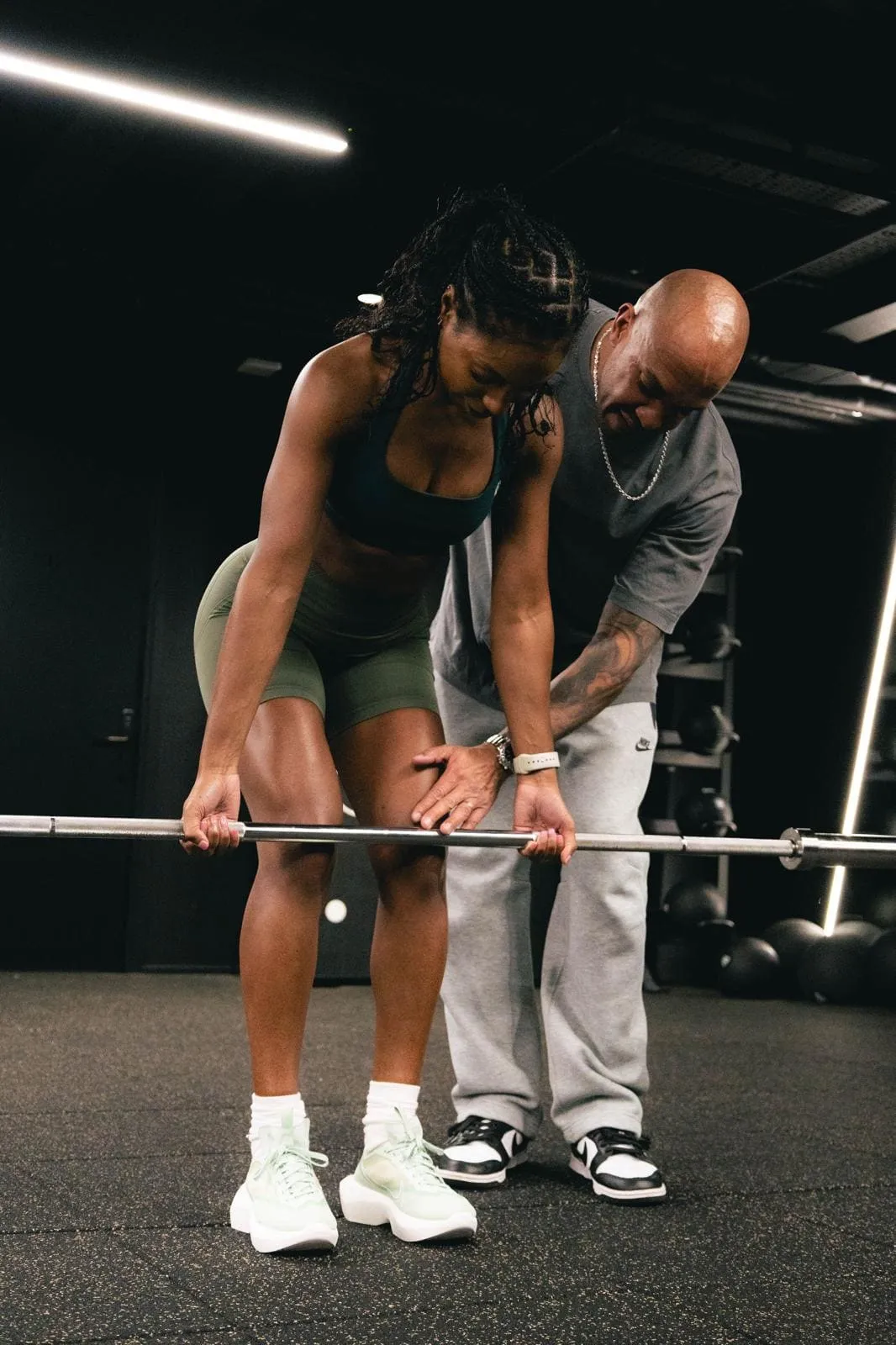 Female athlete lifting barbell with trainer guiding her posture in a gym setting.