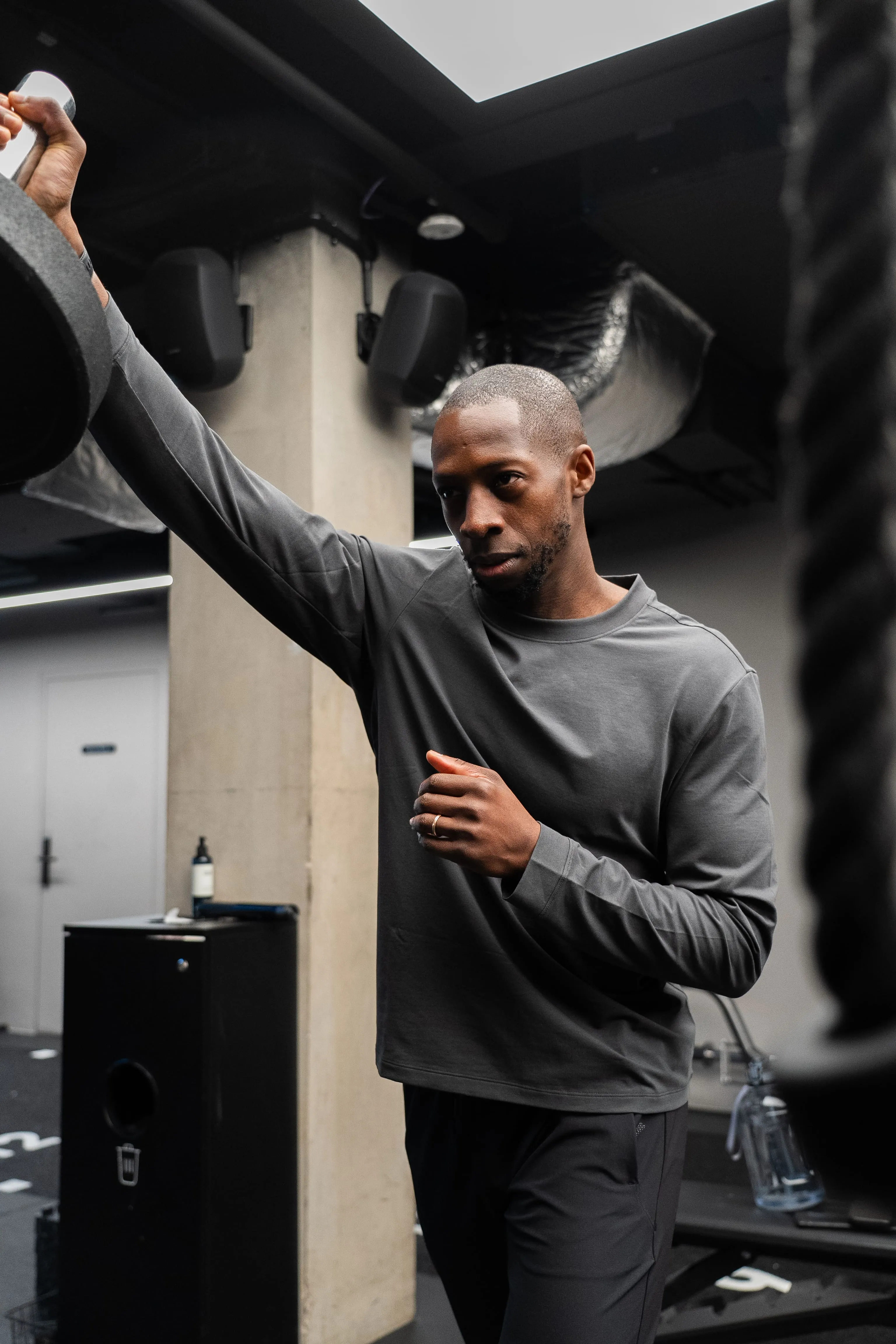 Man in gray long-sleeve shirt resting arm on dumbbell weight in gym.