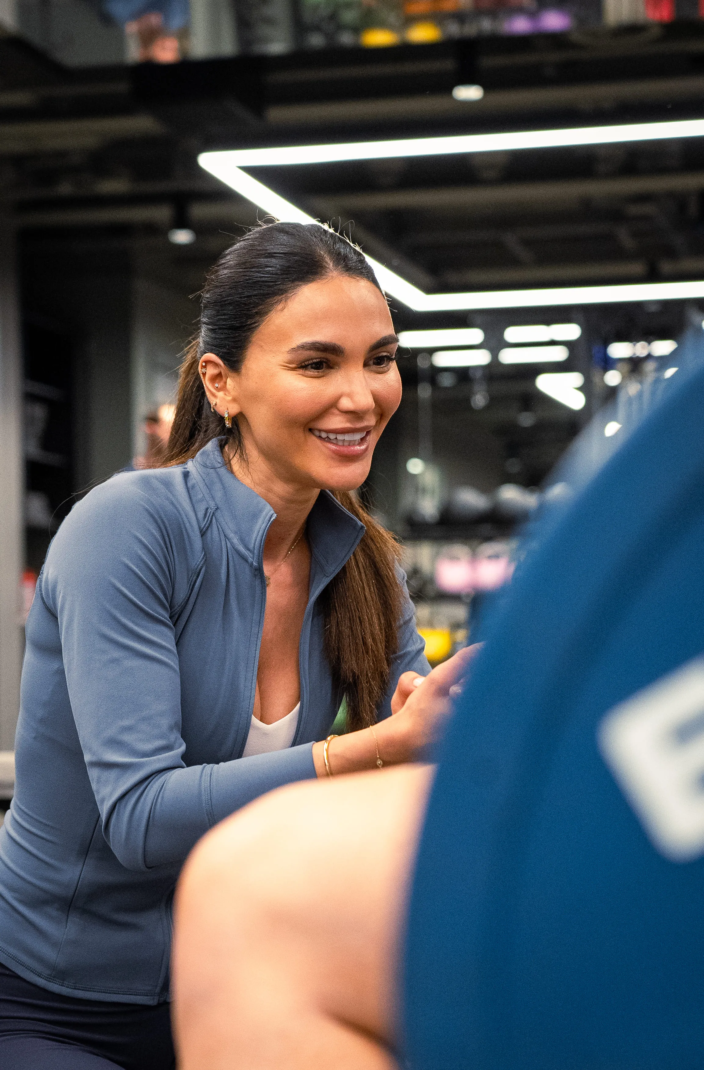 Smiling woman in a blue jacket coaching someone at a gym with modern lighting.