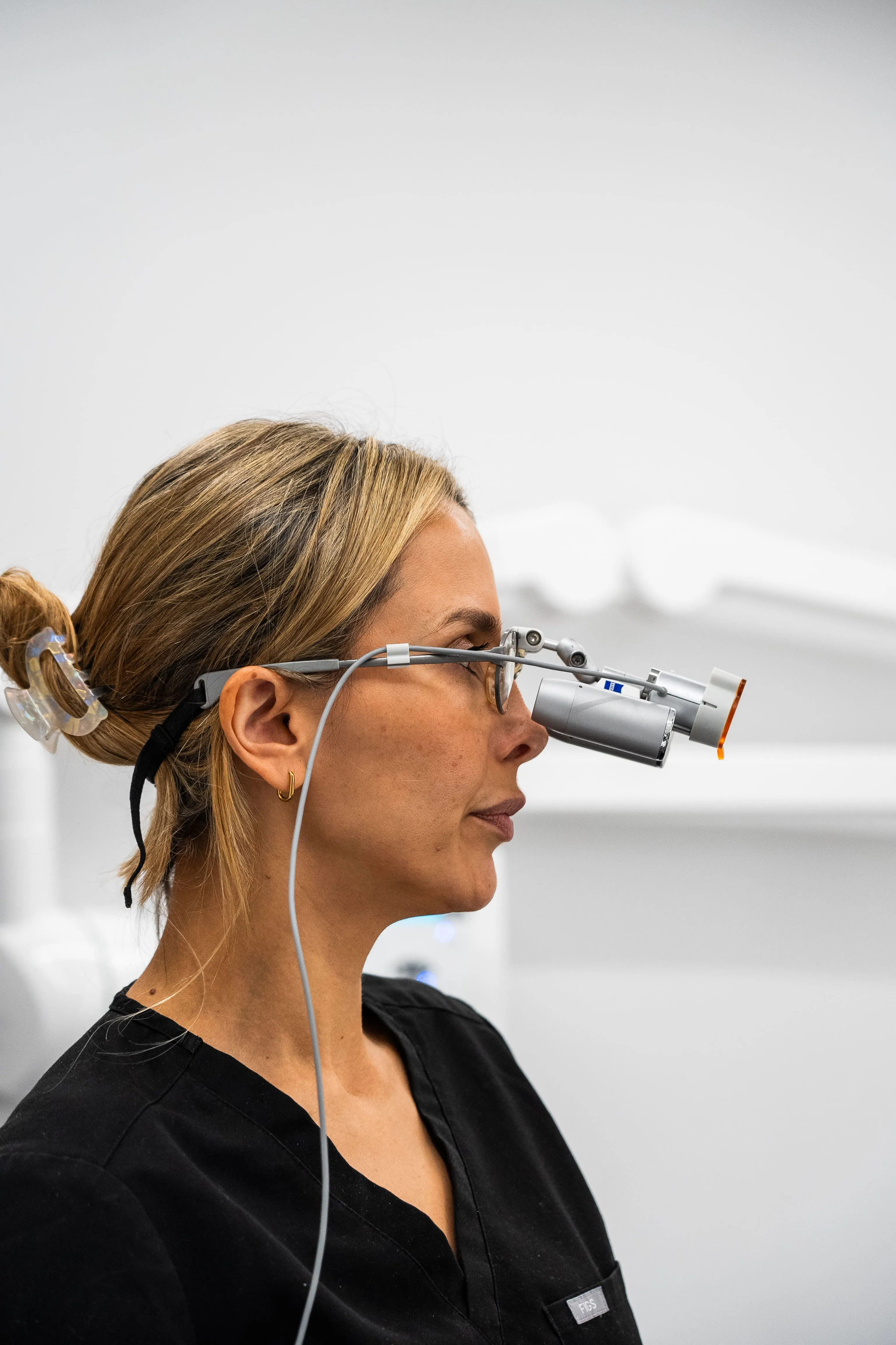 Woman in black scrubs wearing magnifying loupes with light, hair tied back with clip.