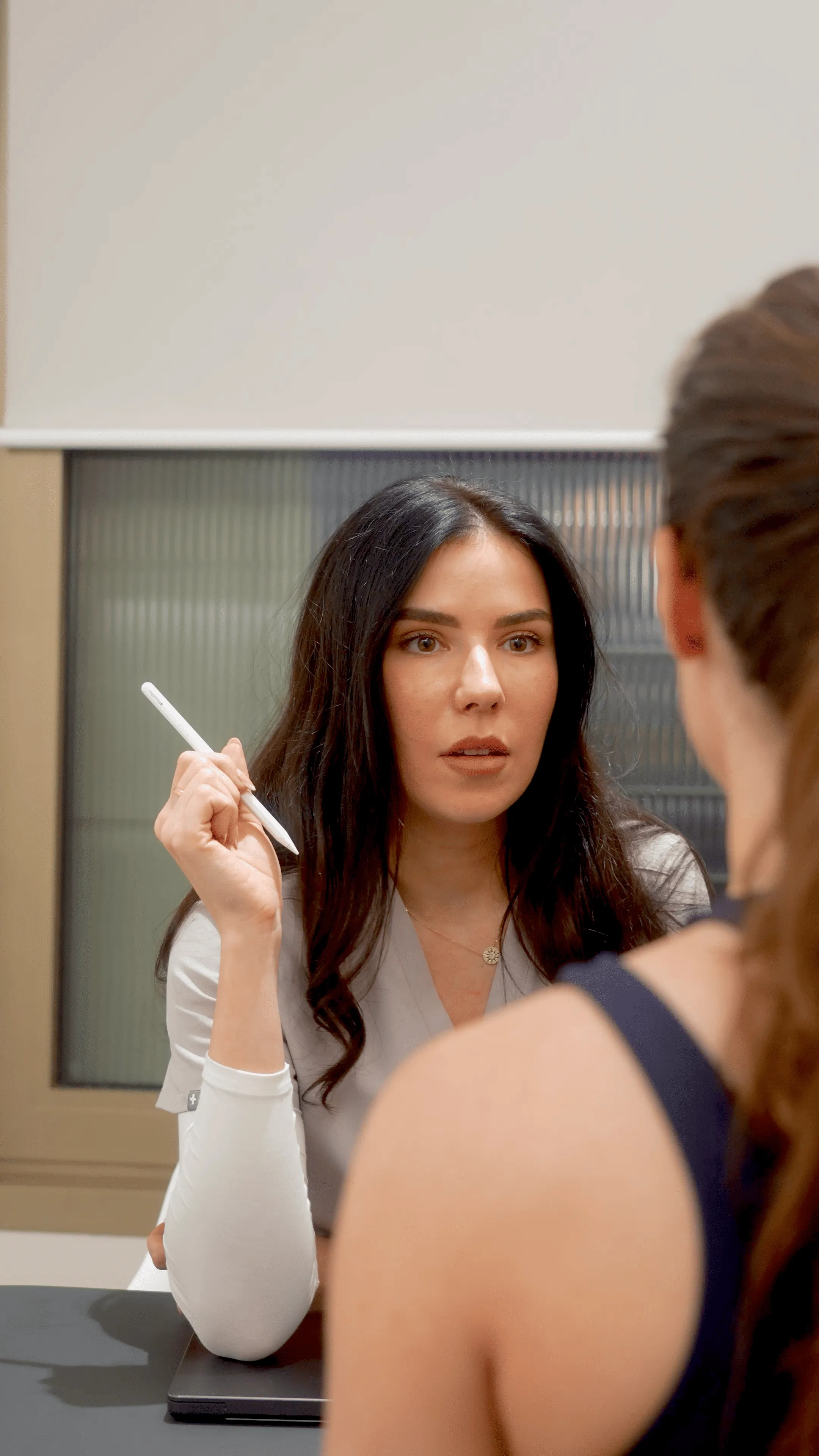 Woman in gray medical scrubs holding a pen, attentively listening to another person.