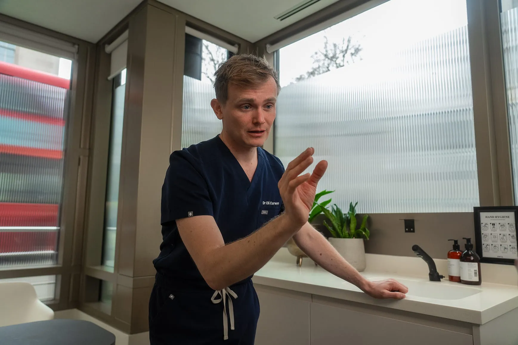 Man in navy medical scrubs gesturing with hand in a modern office with plants and hand hygiene sign.