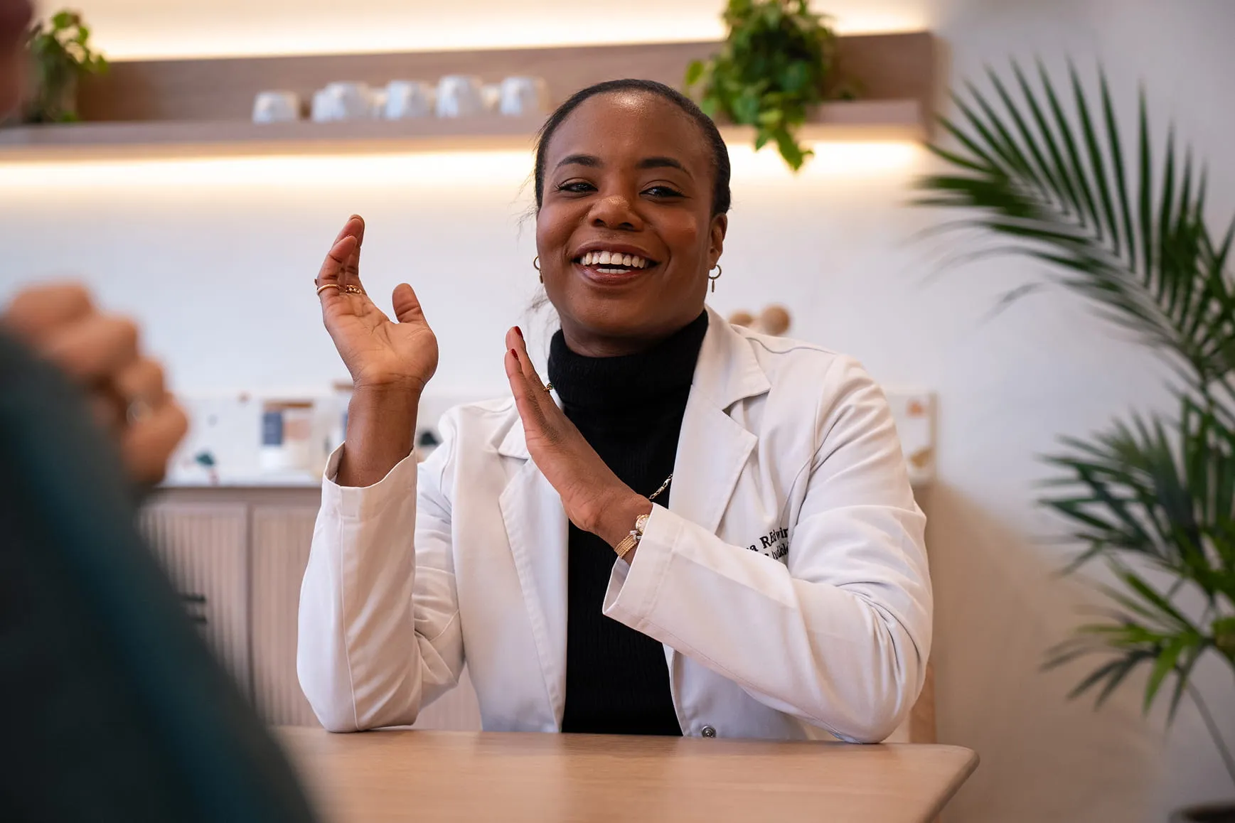 Smiling woman in a white lab coat gestures while talking at a table in a well-lit room with plants.
