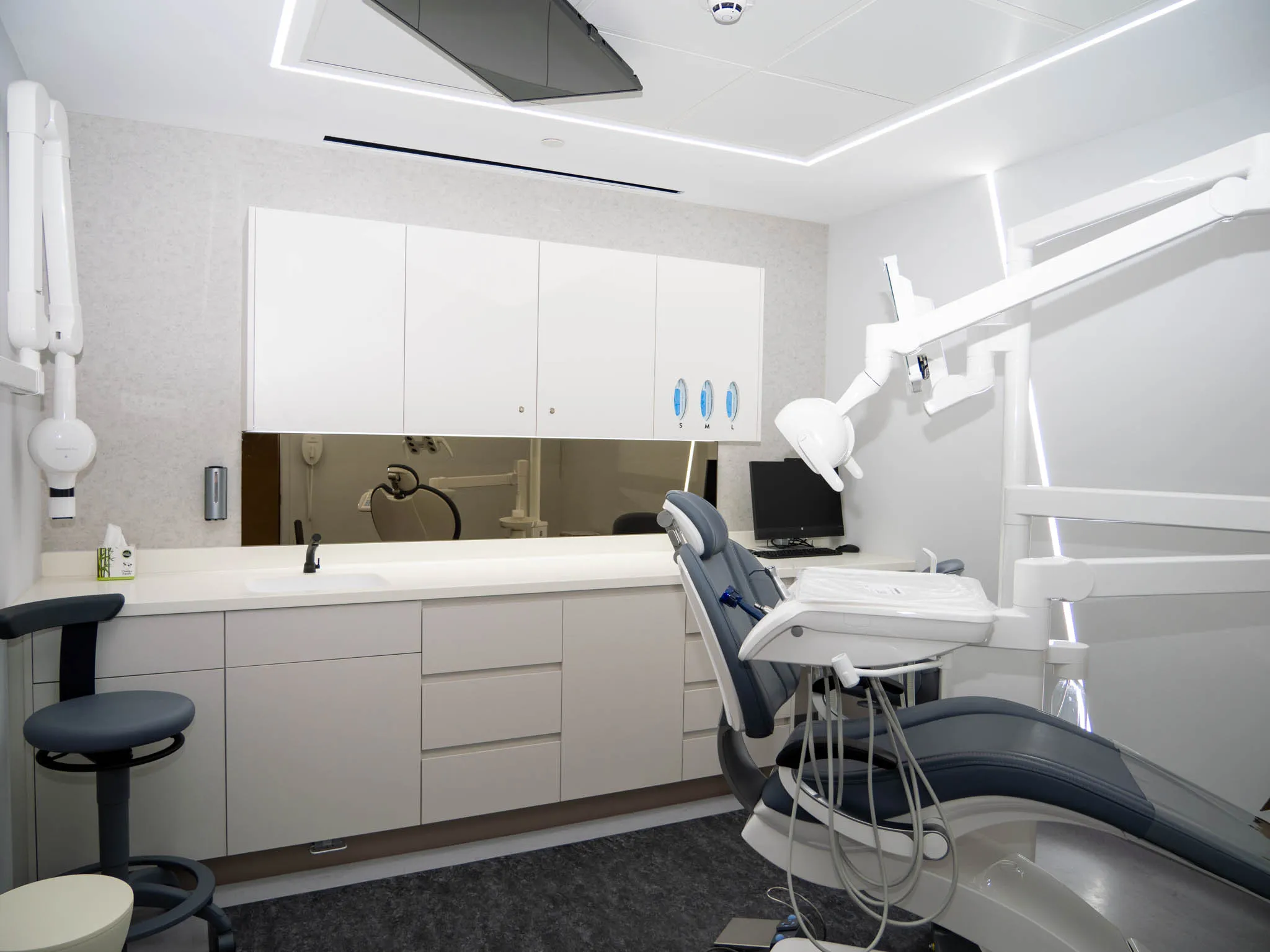 Modern dental clinic room with dental chair, overhead light, cabinets, and computer monitor.