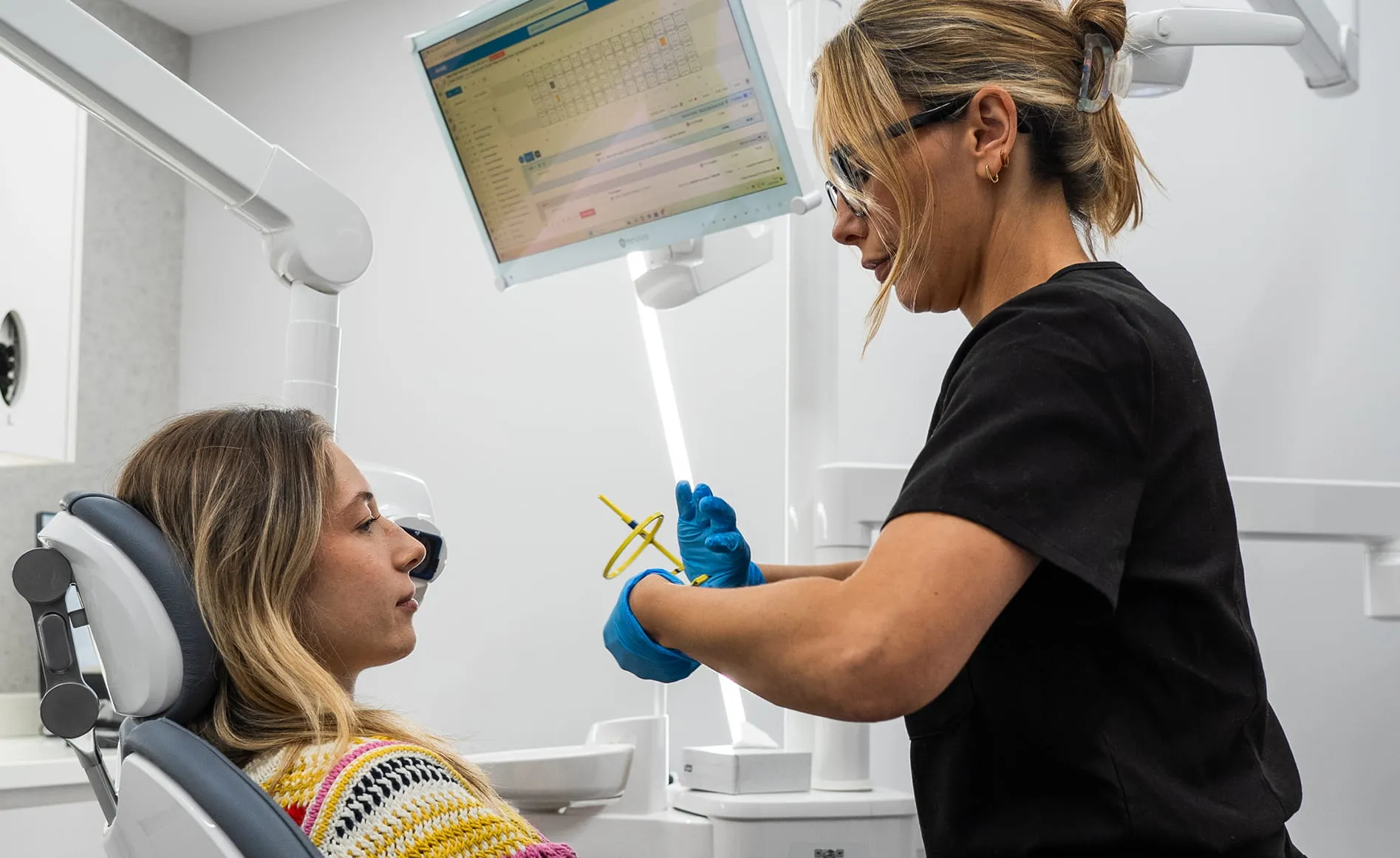 Dentist in black scrubs and gloves preparing tools for a female patient in dental chair.