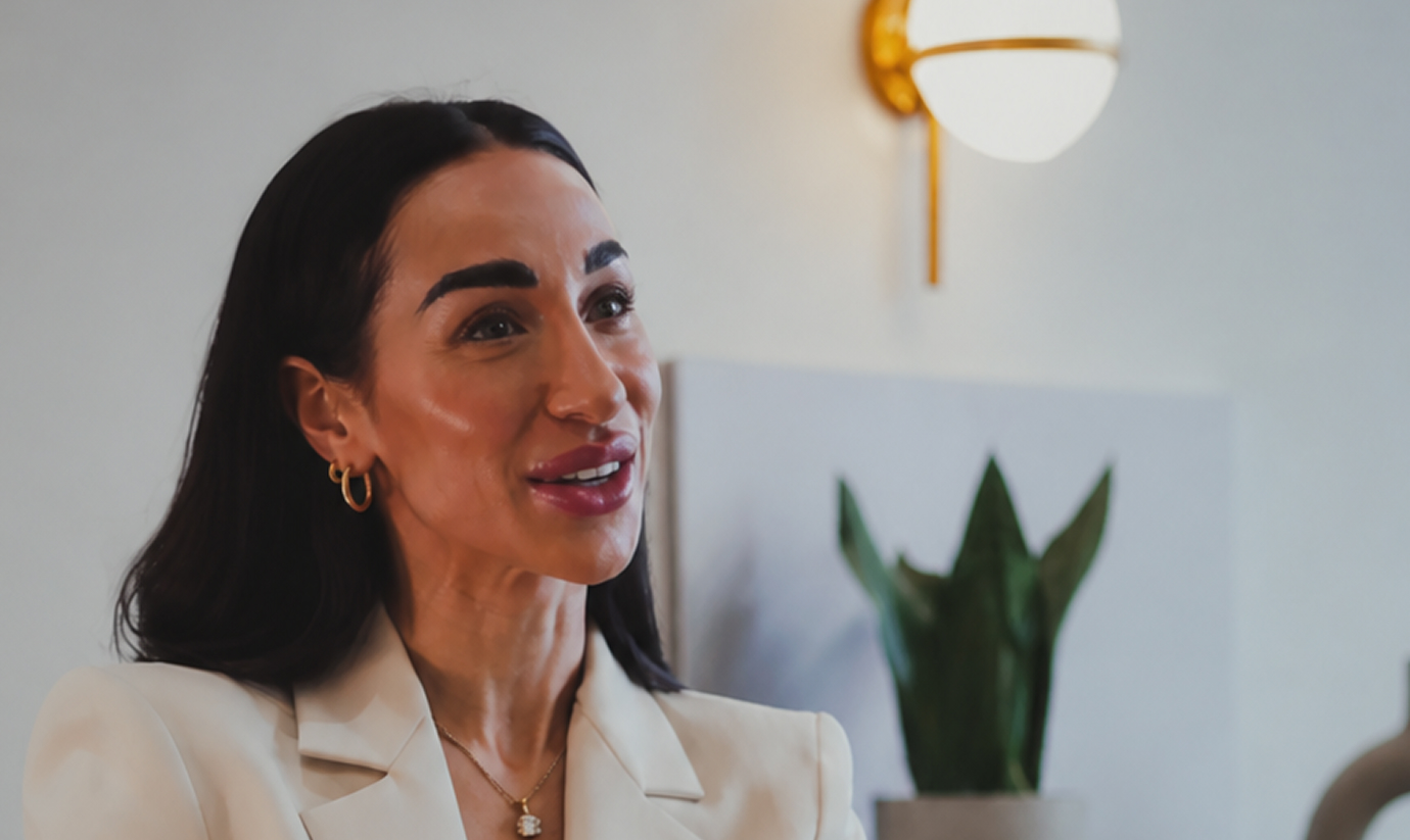 Smiling woman in a white lab coat gestures while talking at a table in a well-lit room with plants.