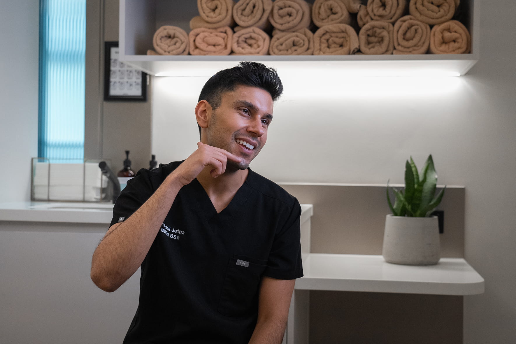 Smiling man in black medical scrubs points to his chin in a clean, modern clinic room.