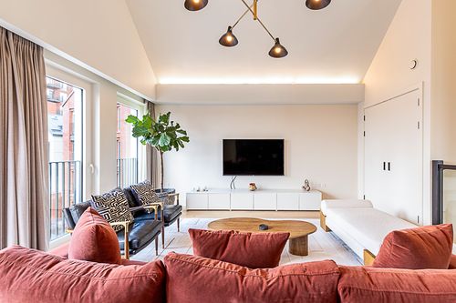 Modern living room with a red sofa, two black armchairs with patterned cushions, a wooden coffee table, white daybed, large plant, and wall-mounted TV.