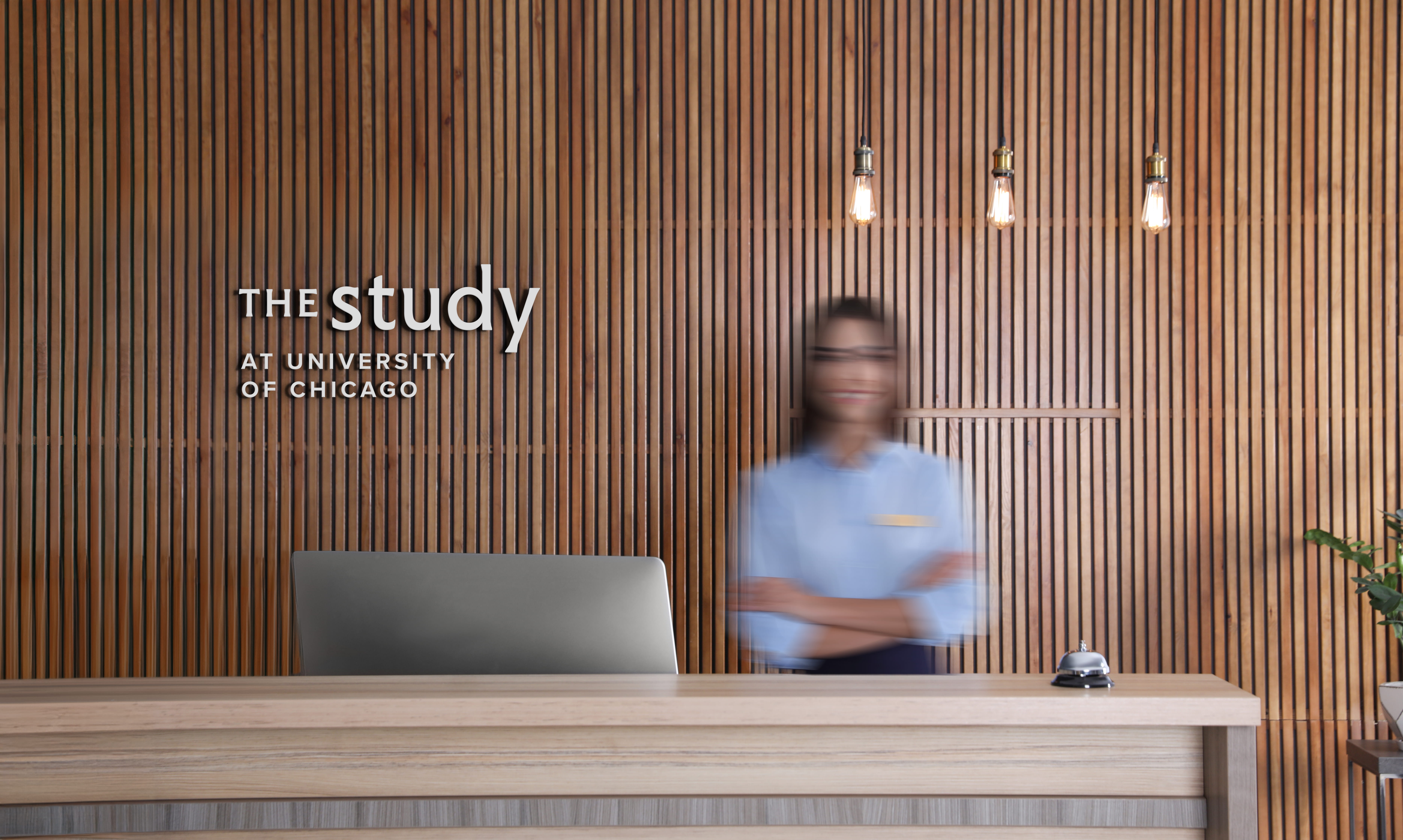 Woman standing at hotel reception desk in front of wood slat wall with Study Hotel branding under edison lightbulbs