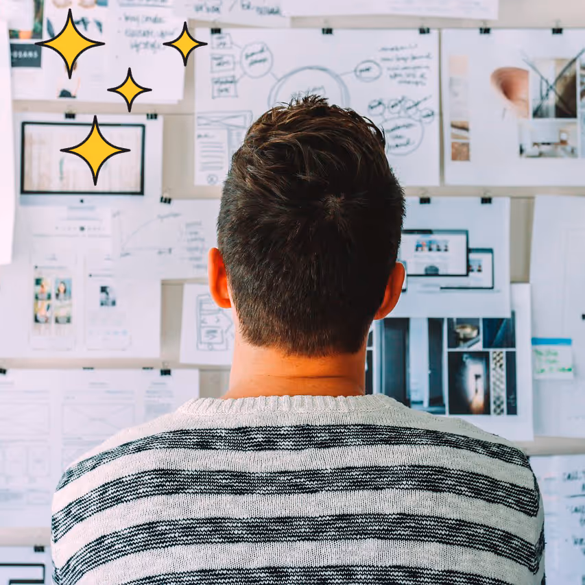 Man with short hair and striped sweater looking at a wall covered with papers and sketches.