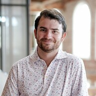 Smiling man with short brown hair and beard wearing a white floral shirt in a bright indoor setting.